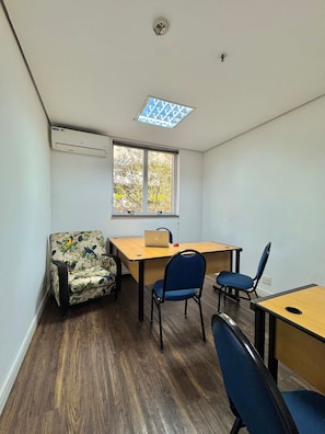 A small, well-lit room with a wooden desk, two chairs, a patterned armchair, a window with a view of trees, and an air conditioning unit.