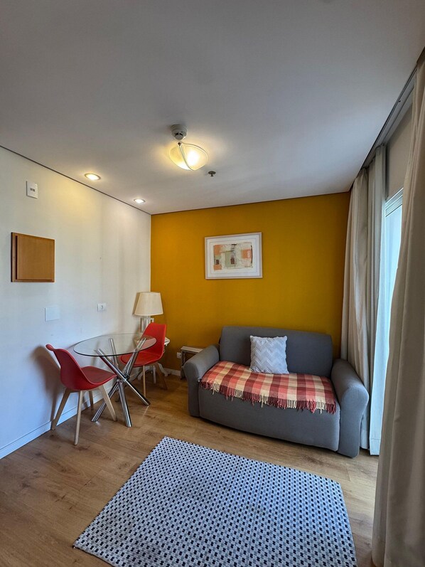 Un pequeño espacio de sala con un sofá, una silla roja, una mesa de vidrio y una lámpara.