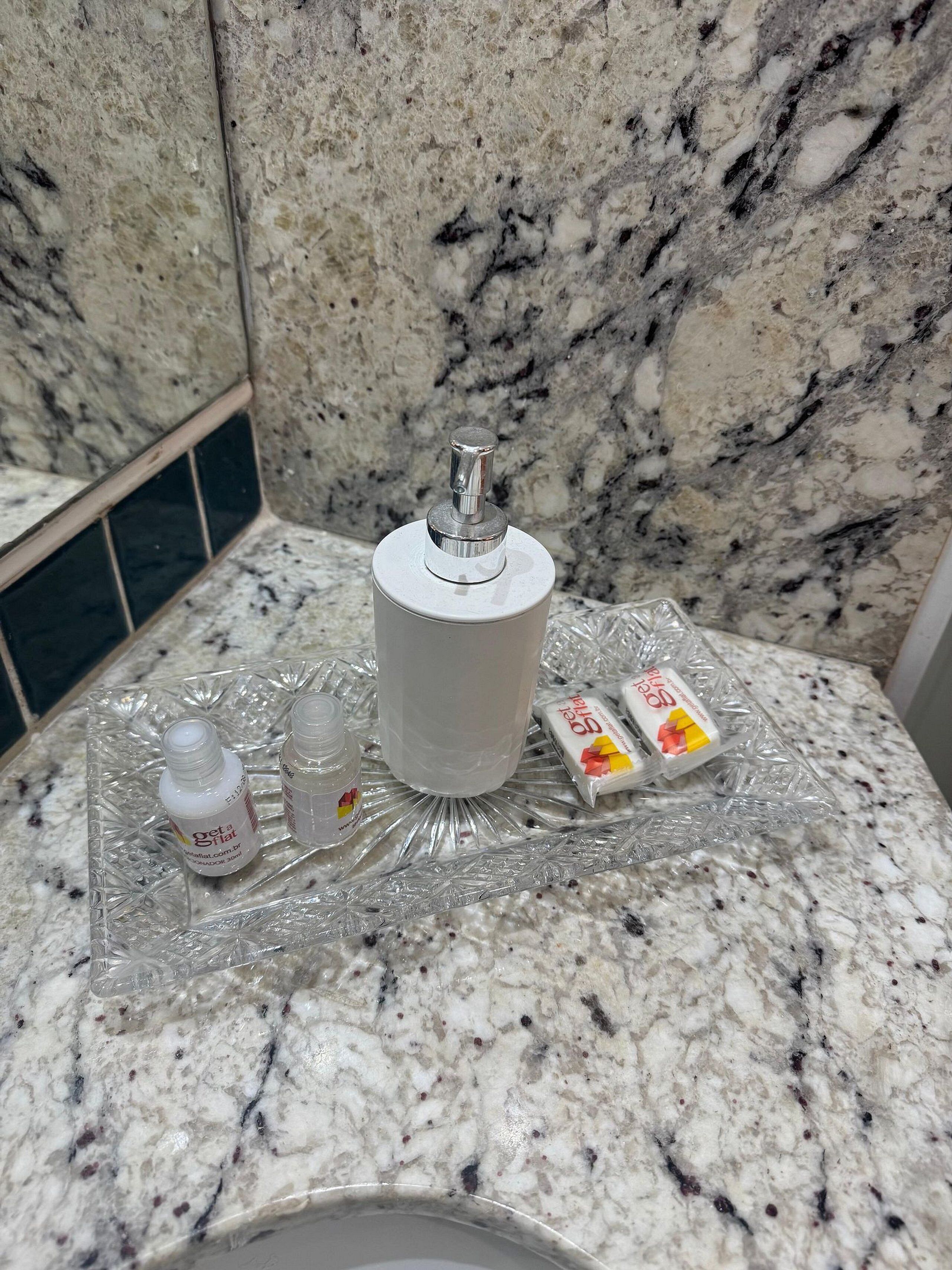 Un baño con encimera de mármol, dispensador de papel higiénico blanco y artículos de aseo en una bandeja de vidrio.