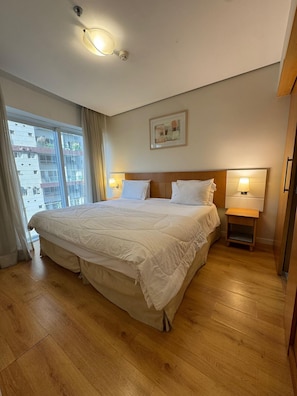 A hotel bedroom with a large bed, two bedside lamps, a ceiling light, a window with curtains, and a city view. - Get a Flat 601 Ibirapuera -Jardins (São Paulo)