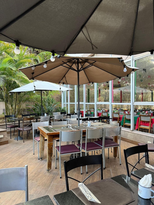 An outdoor cafe with tables and chairs under umbrellas, surrounded by large windows and greenery.