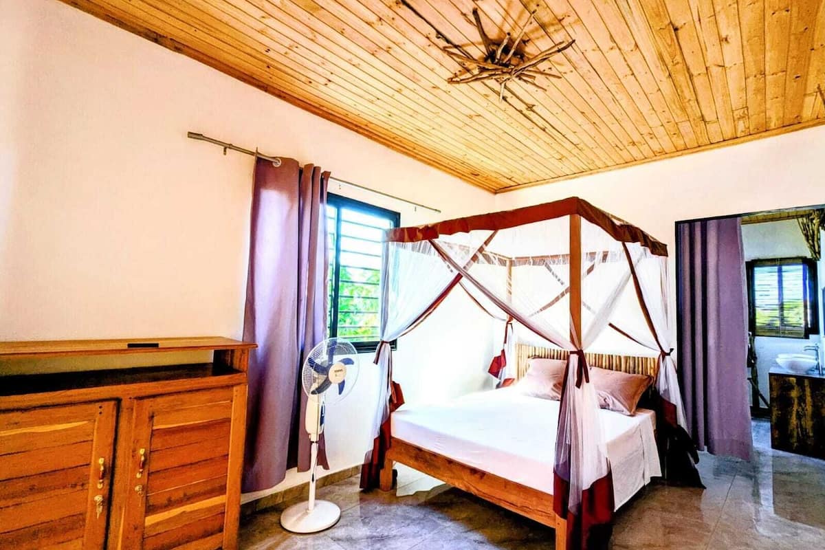 Un dormitorio con techo de madera, cama con dosel, ventilador en la mesita de noche y una ventana con cortinas.