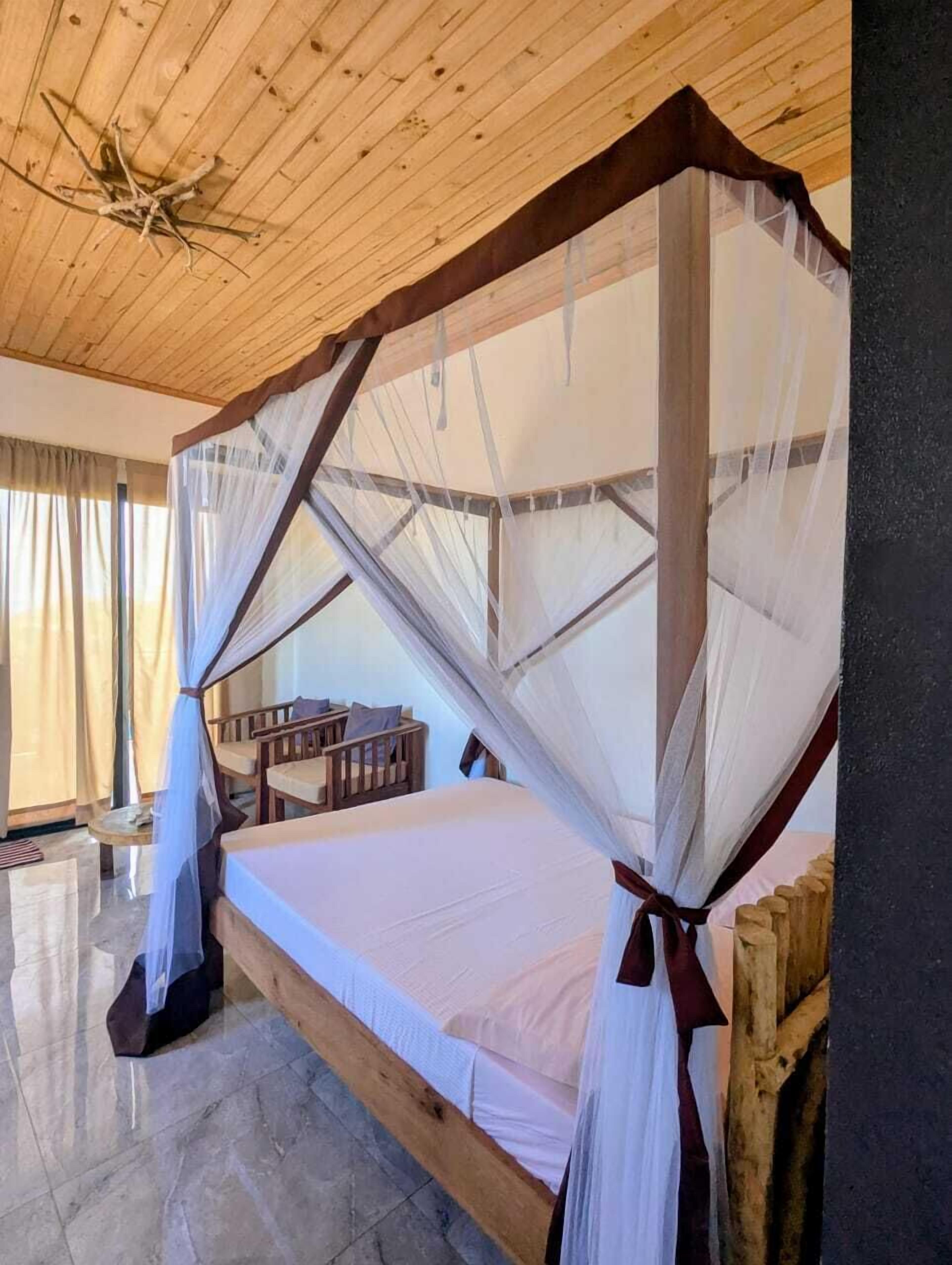 A wooden-framed bed with a canopy, a small wooden table, and a chair in a room with a tiled floor and wooden ceiling.