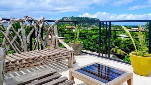 Un banco de madera y una mesita en un balcón con vistas a zonas verdes y una colina distante.