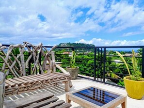 Eine hölzerne Bank und ein kleiner Tisch auf einem Balkon mit Blick auf Grünflächen und eine ferne Hügelkette.