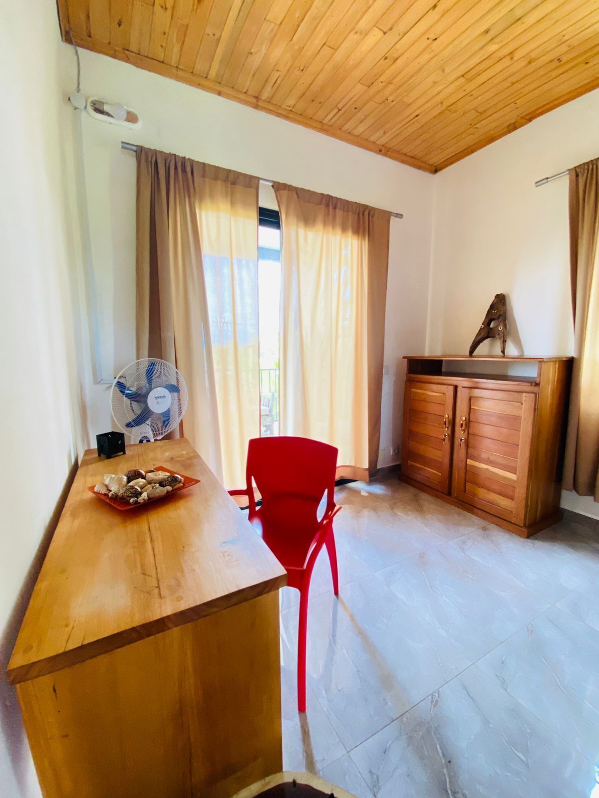 Une pièce avec un plafond en bois, une table en bois avec une chaise rouge, un ventilateur et un meuble sur lequel est posé un objet décoratif.