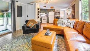 A modern living room with a sofa, ottoman, and armchair, a kitchen in the background, and a large window with curtains. - Peaceful Brookside Getaway ~10 Mi to Okemo Slopes! (Chester)