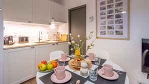 Cozinha moderna com armários brancos, uma mesa de jantar posta com pratos e xícaras cor-de-rosa, e uma colagem de fotos emolduradas na parede.