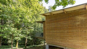 Una cabaña de madera con terraza metálica, rodeada de un denso bosque.