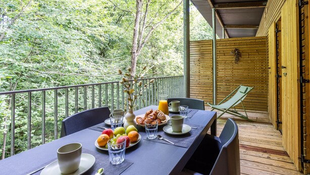 Mesa dispuesta para el desayuno, con frutas, croissants y bebidas, en un balcón con vistas a un área boscosa.
