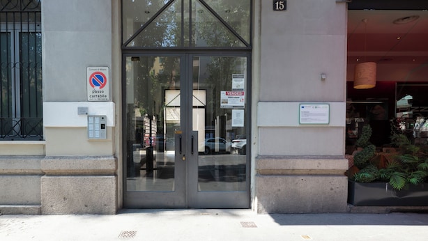 Une entrée d’immeuble avec une porte en verre, un panneau indiquant qu’il est interdit de se garer et le chiffre « 15 » peint sur le mur.