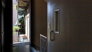 Un couloir étroit avec une porte sur la droite, une rambarde en métal et un petit balcon agrémenté de plantes en pot.