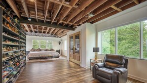 Een ruime kamer met een groot houten plafond, een bed met uitzicht op de omgeving, een lederen fauteuil en een schoenenverzameling op planken.
