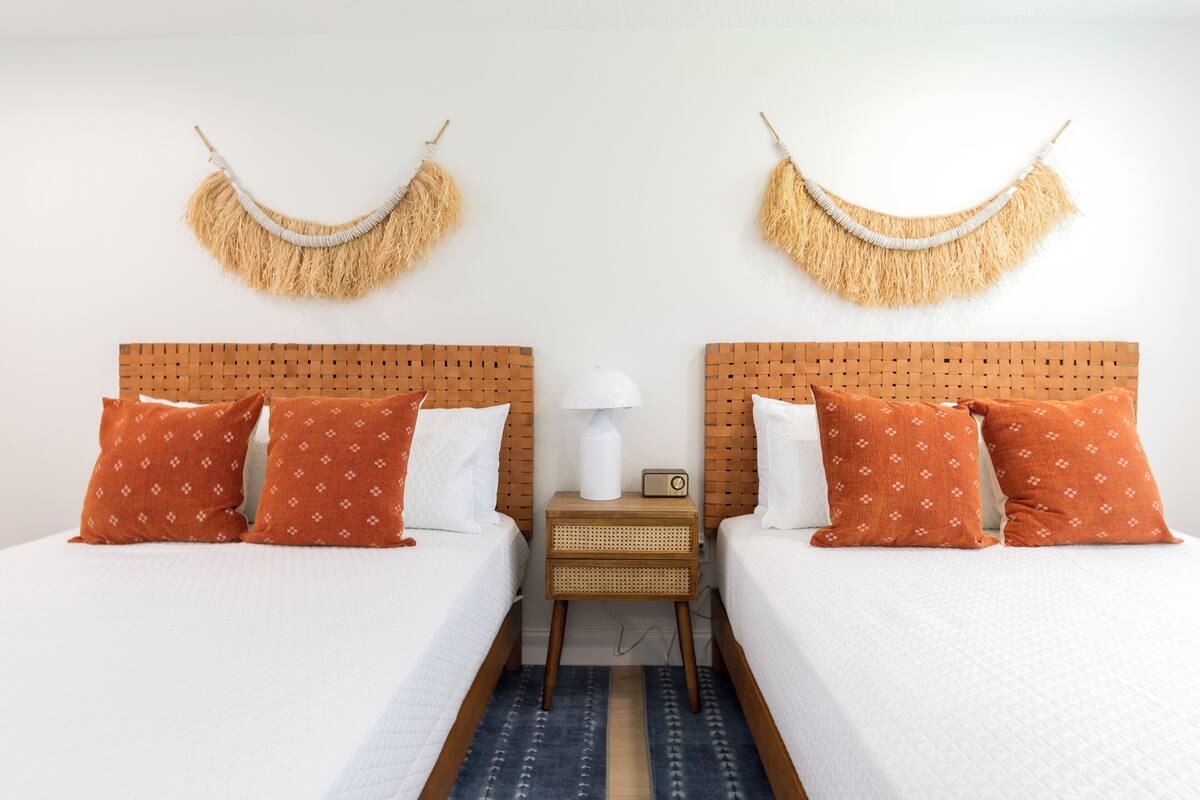 A hotel room with two beds, each with a wooden headboard, white bedding, and orange patterned pillows. There is a small bedside table with a lamp and a radio.