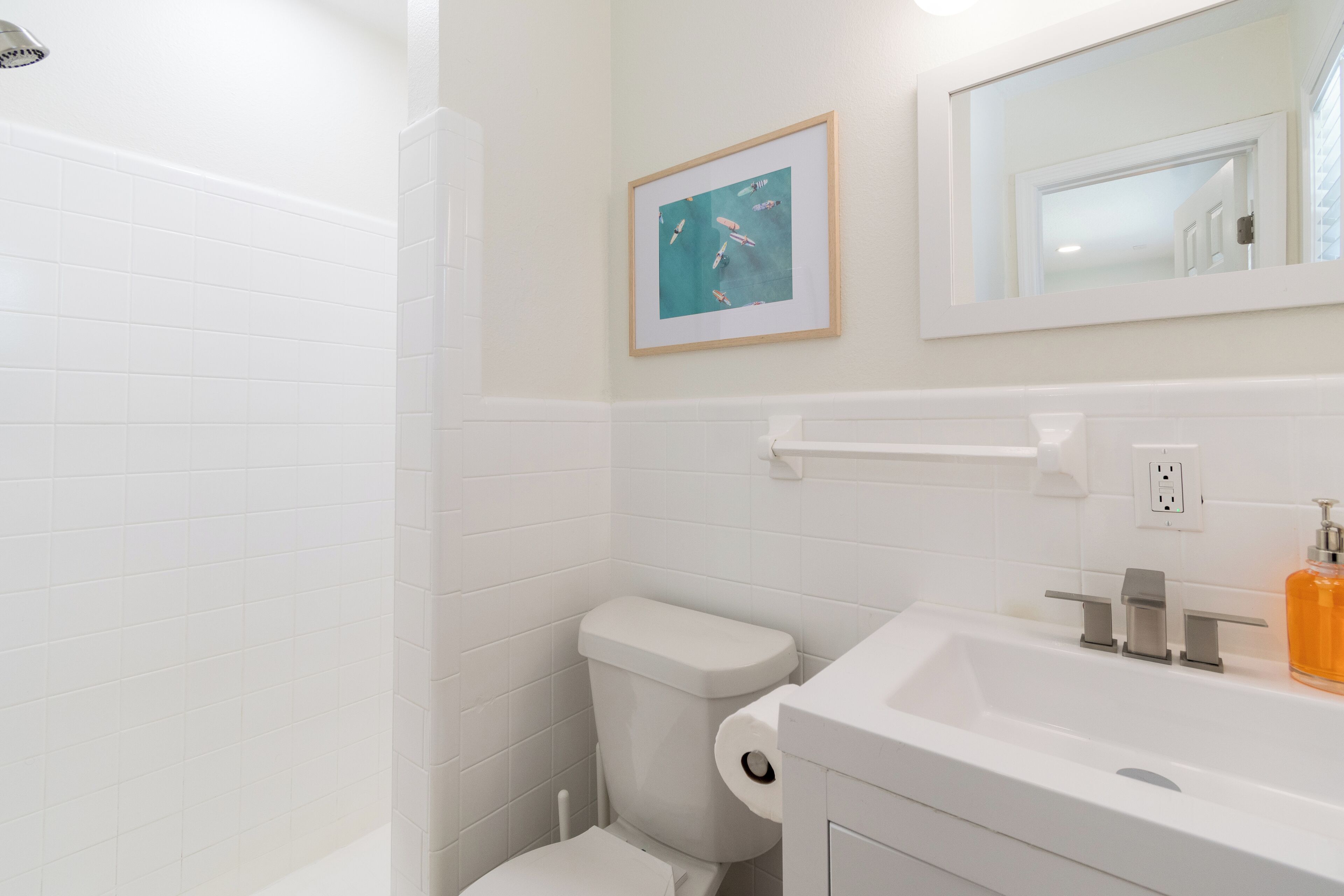 A bathroom with white tiles, a toilet, a sink with a soap dispenser, and a framed picture on the wall.