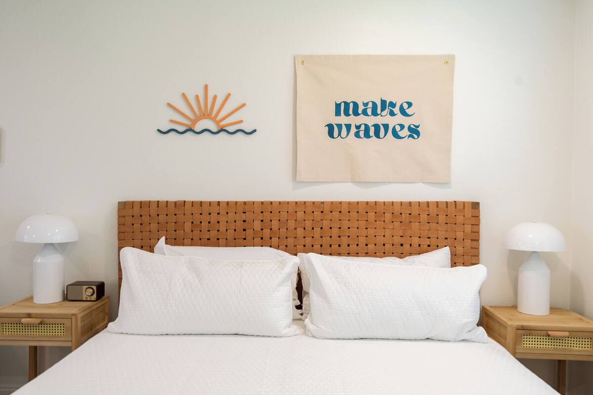 A neatly made bed with white bedding and two pillows, a wooden headboard, and a wall with a sun and wave decoration and a 'make waves' sign.