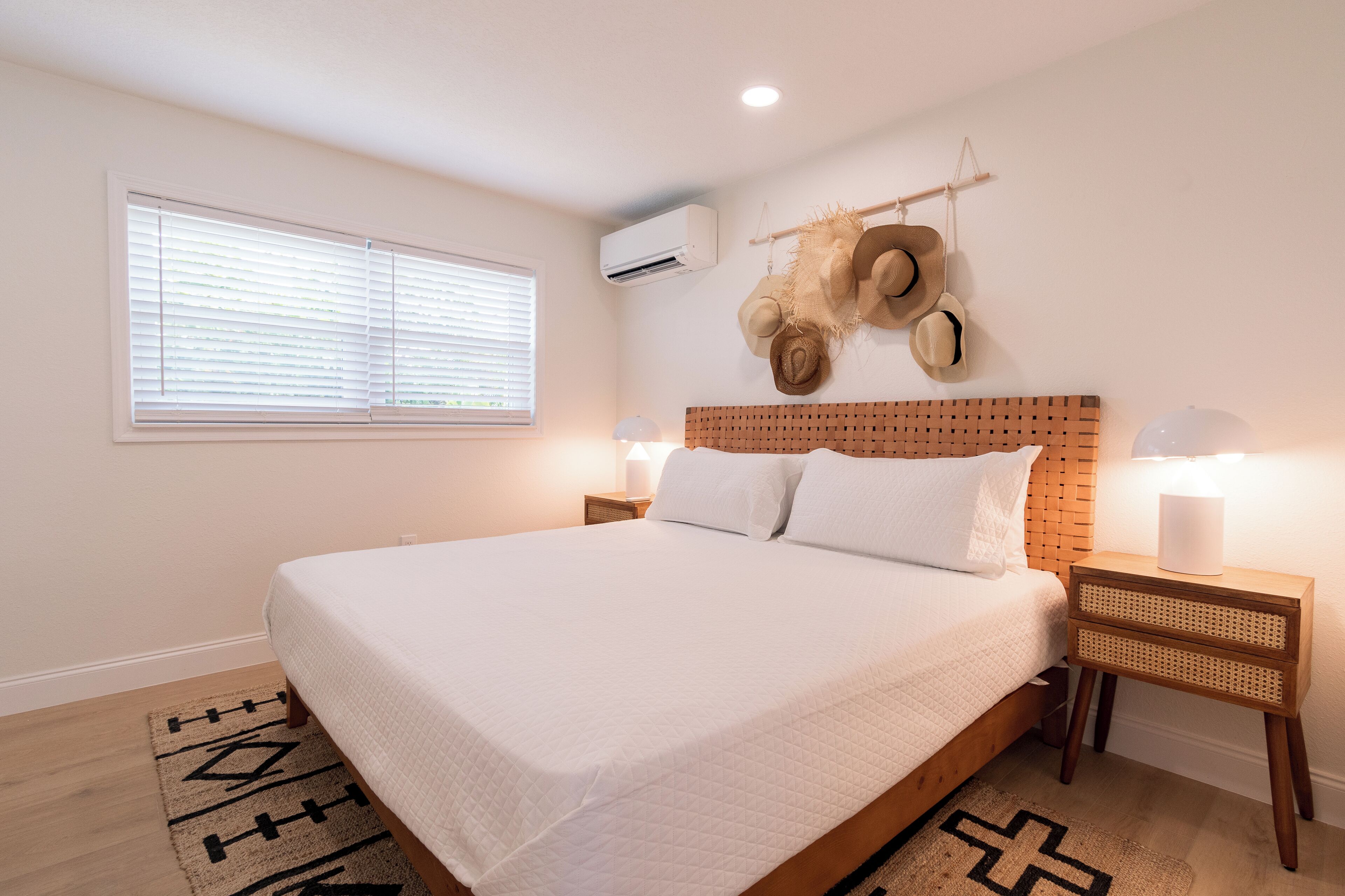 A neatly arranged bedroom with a large bed, a headboard with a woven pattern, two bedside tables with lamps, and a wall-mounted air conditioner.