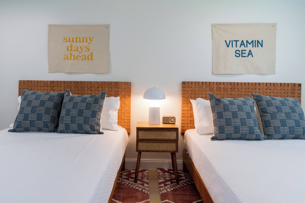 A double bed room with a wooden headboard, a bedside table with a lamp, and two wall signs reading "sunny days ahead" and "VITAMIN SEA".