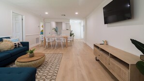 A modern living room with a sofa, coffee table, TV, and dining area. - The Juni (Cocoa Beach)