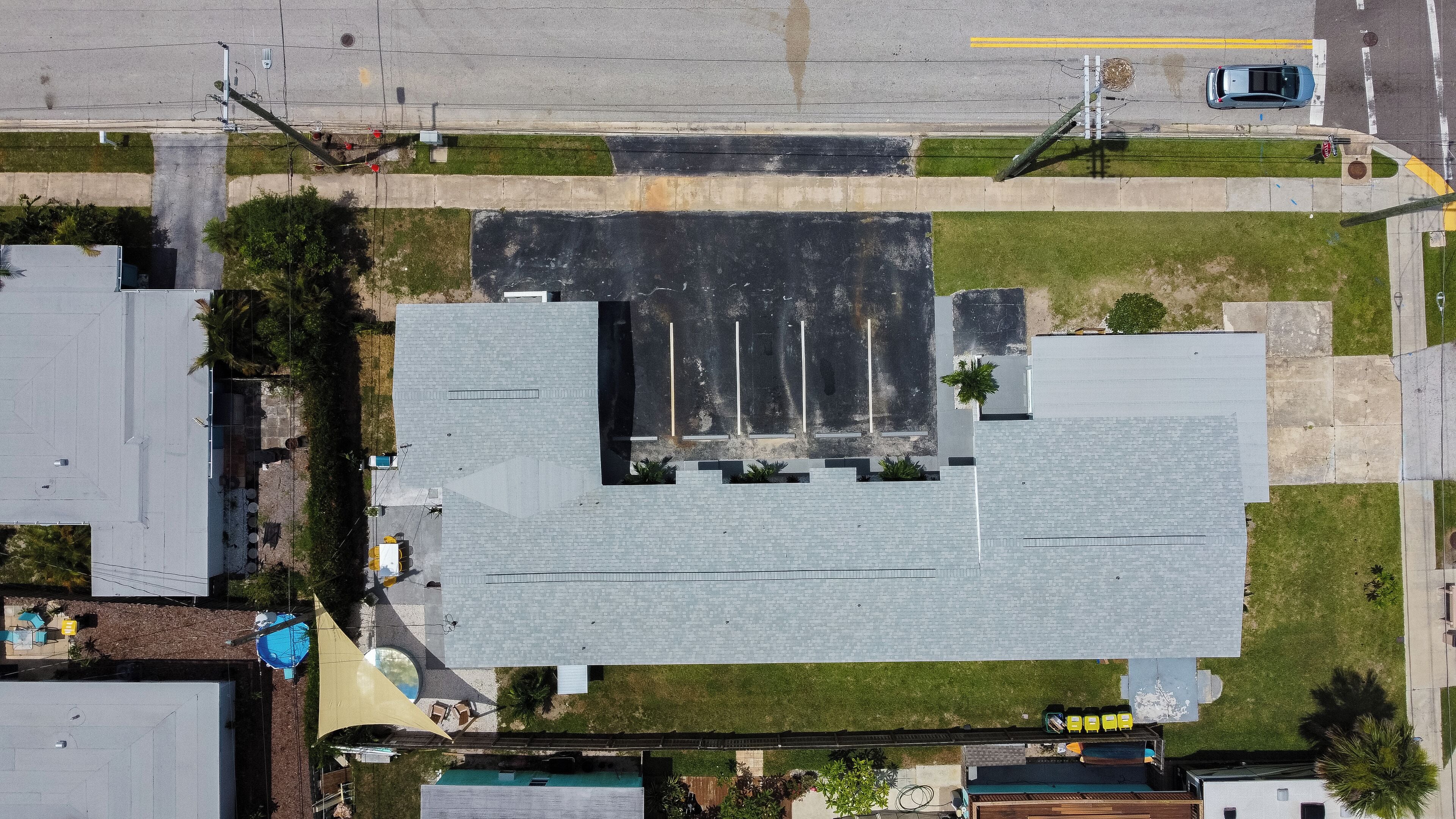 An aerial view of a parking lot with multiple rows of parking spaces, a building with a flat roof, and a grassy area with a pool and outdoor seating.