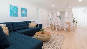 A modern living room with a sectional sofa, a dining area with white chairs, and a kitchen in the background. - The Juni (Cocoa Beach)