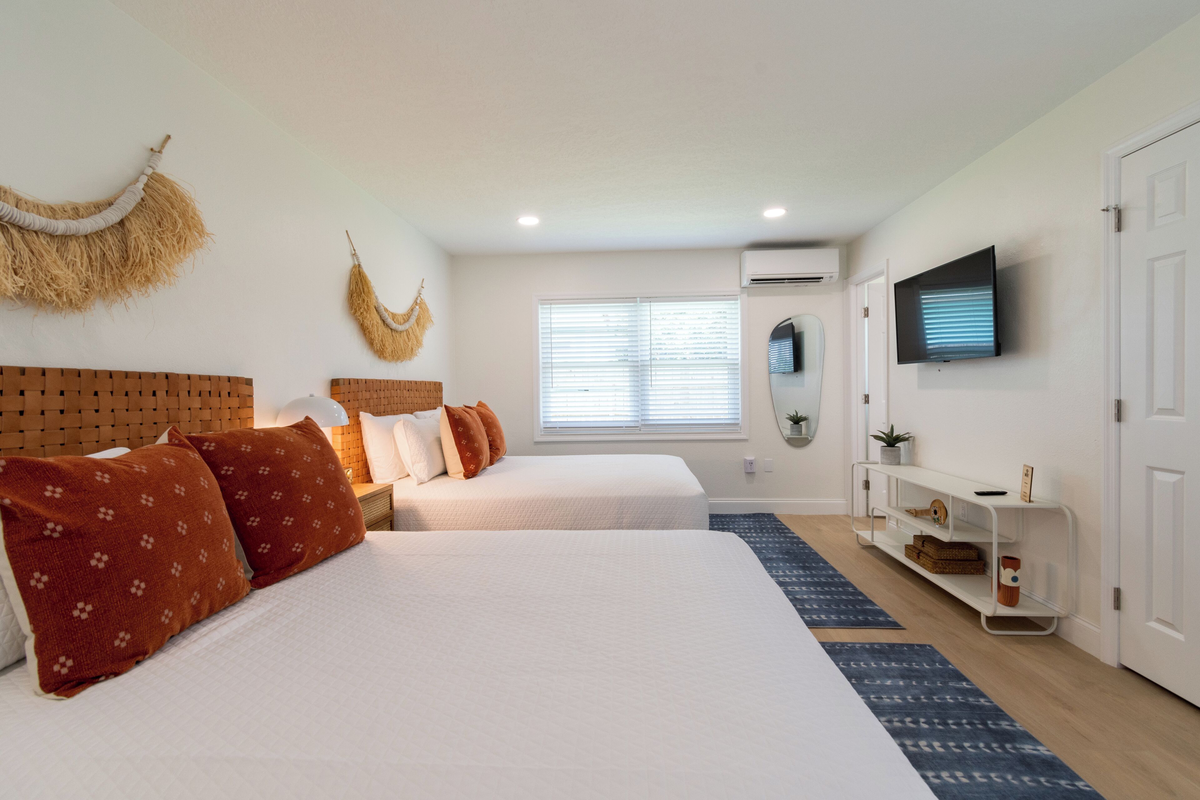 A hotel room with two beds, a TV, a window with blinds, a wall-mounted air conditioner, and a small shelf with decorative items.