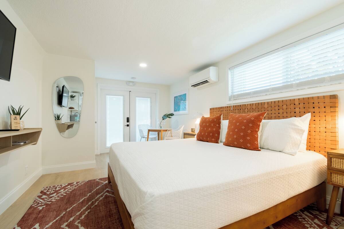 A neatly arranged bedroom with a large bed, a bedside table, a mirror, and a window with blinds.