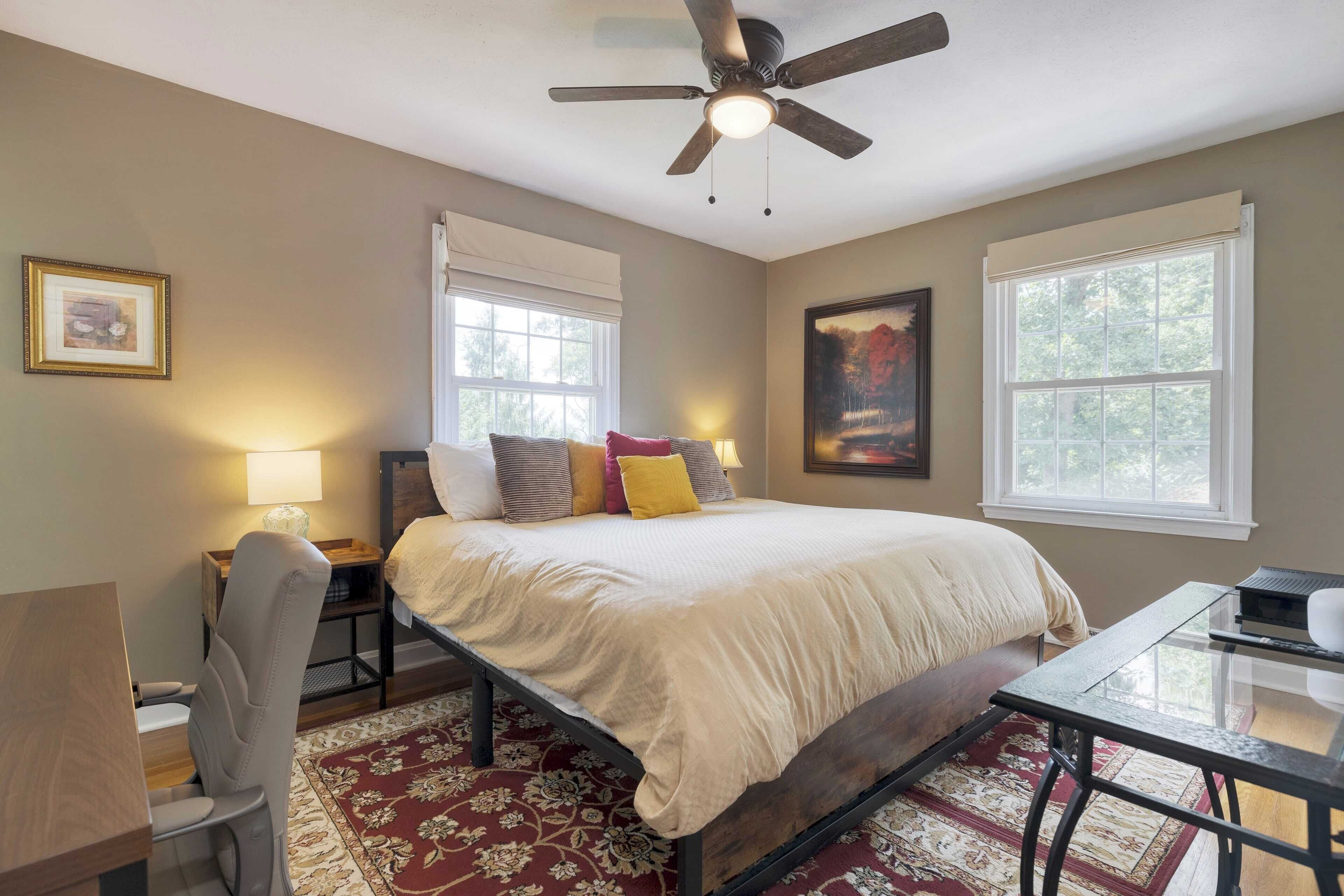 A bedroom with a bed, a desk, a chair, a ceiling fan, and two windows.