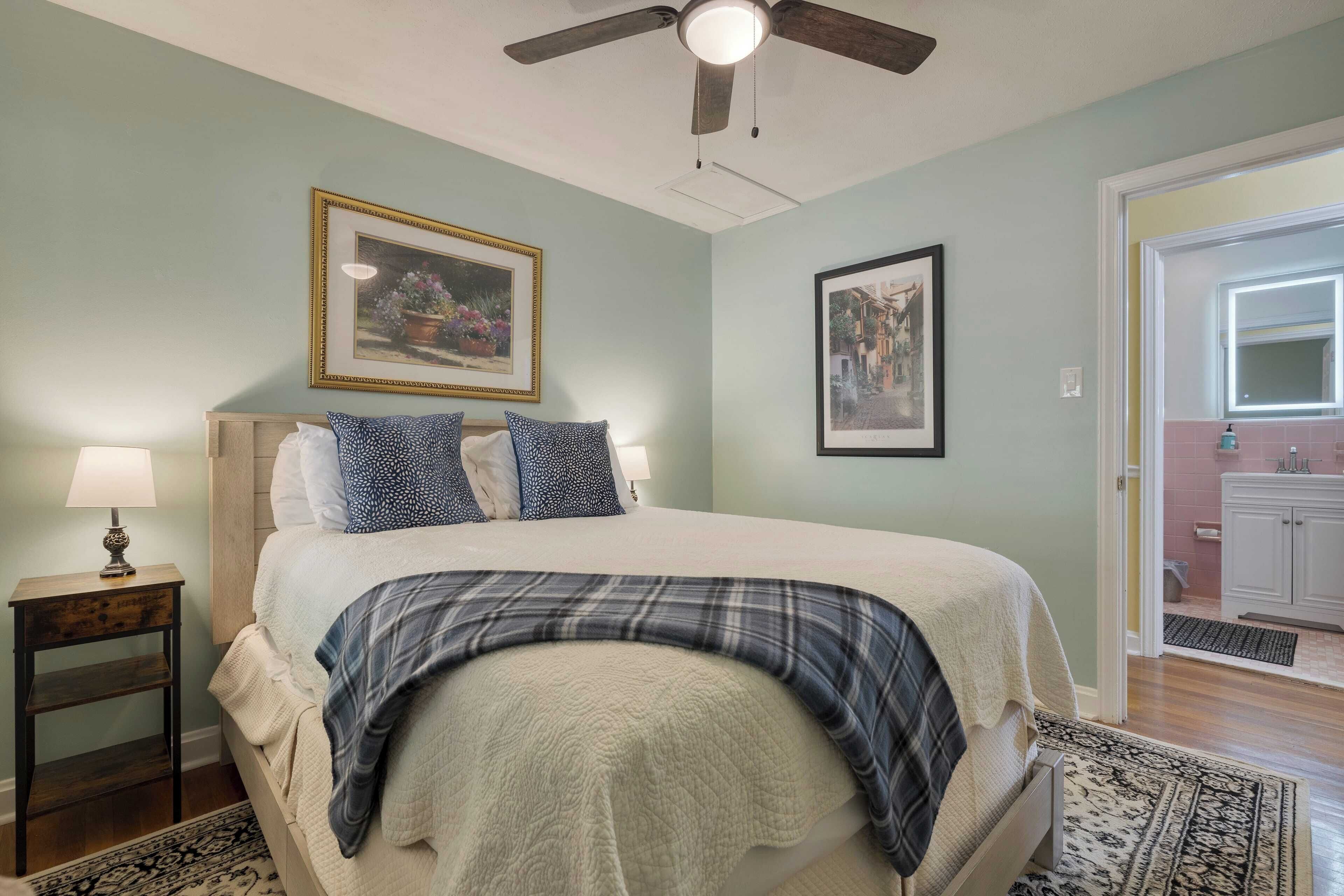 Un dormitorio con cama, mesita de noche, ventilador de techo y puerta que da a un baño.