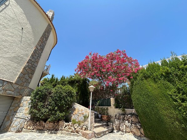 Un edificio de piedra con fachada curva, un árbol de flores rosadas y un jardín bien cuidado con diversas plantas y una escalera de piedra.