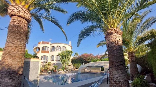 A pool area with a hammock, palm trees, and a building with arched windows in the background. - VILLA LOS ALMENDROS WITH PRIVATE POOL  (Calpe , Alicante)