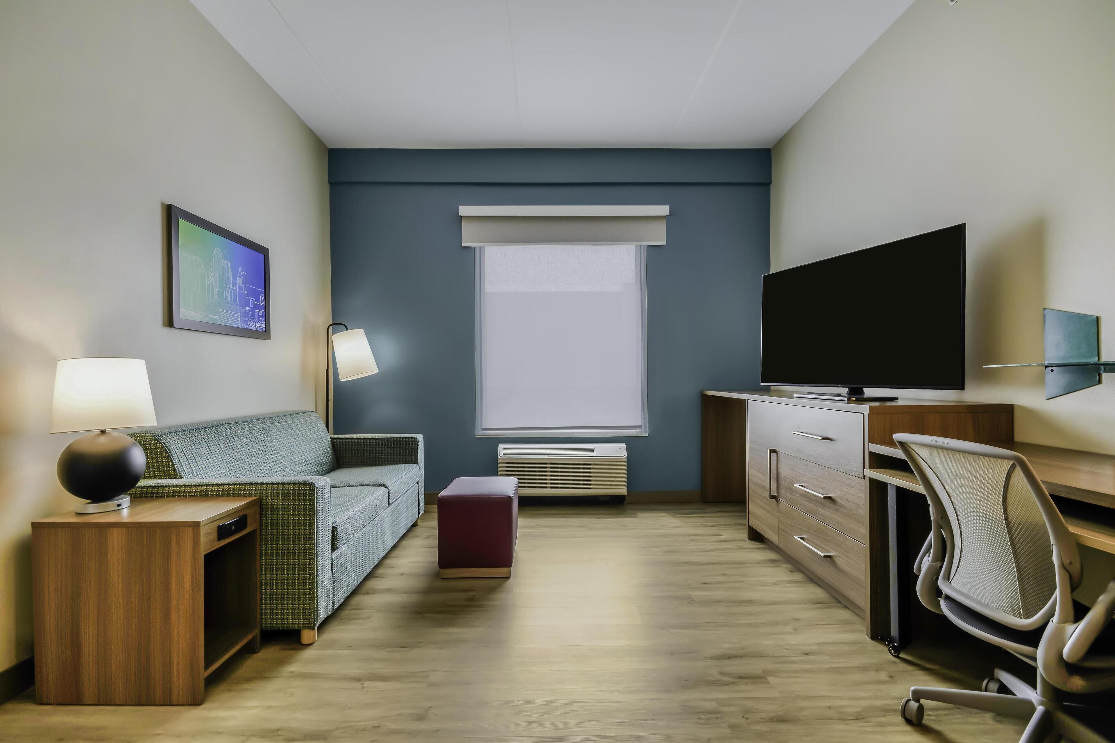 Ett kompakt hotellrum med en soffa, ett litet bord med en lampa, en tv, ett skrivbord med en stol och ett fönster med persienner.