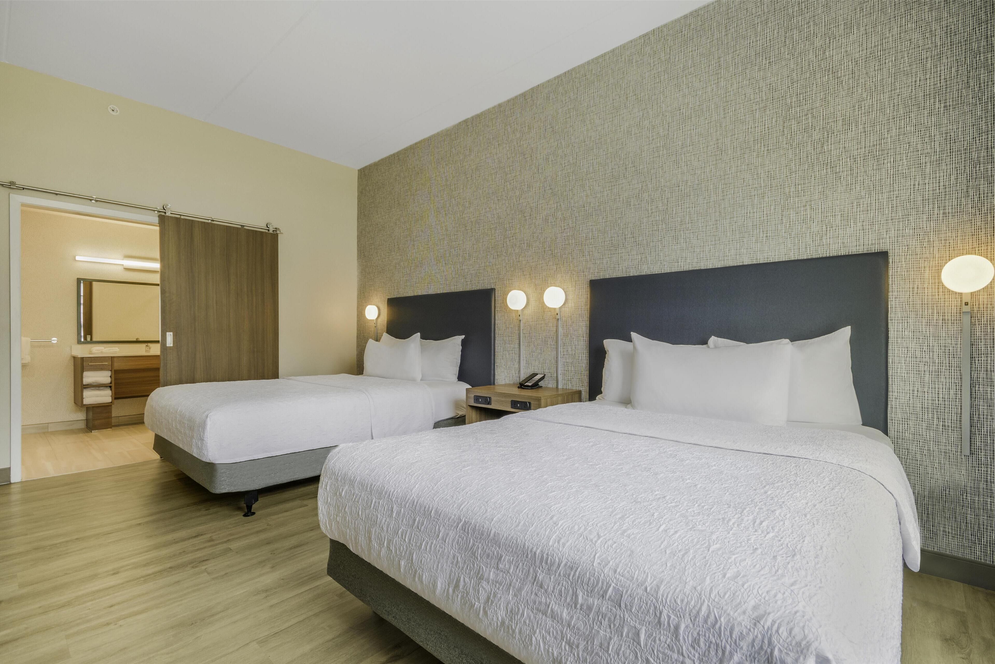 Une chambre d’hôtel avec deux lits, un plancher en bois, un mur à texture et une salle de bain visible à travers une porte ouverte.