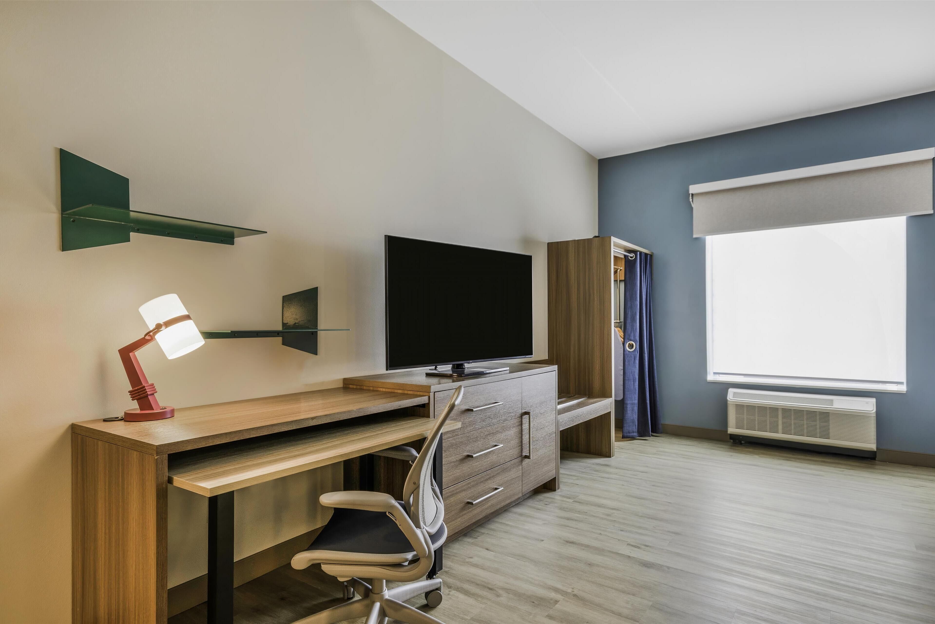 A modern room with a wooden desk, a chair, a flat-screen TV, a lamp, a shelf, and a window with curtains.