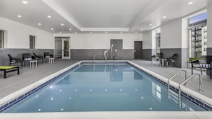 Una piscina interior moderna con agua azul cristalina, rodeada de paredes grises y blancas, y equipada con varias zonas de estar y una escalera que conduce a un nivel superior.