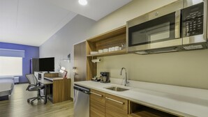 Cocina moderna con microondas incorporado, cafetera y lavamanos. Hay un mueble de madera con estante sobre el lavamanos y un pequeño escritorio con silla y lámpara.