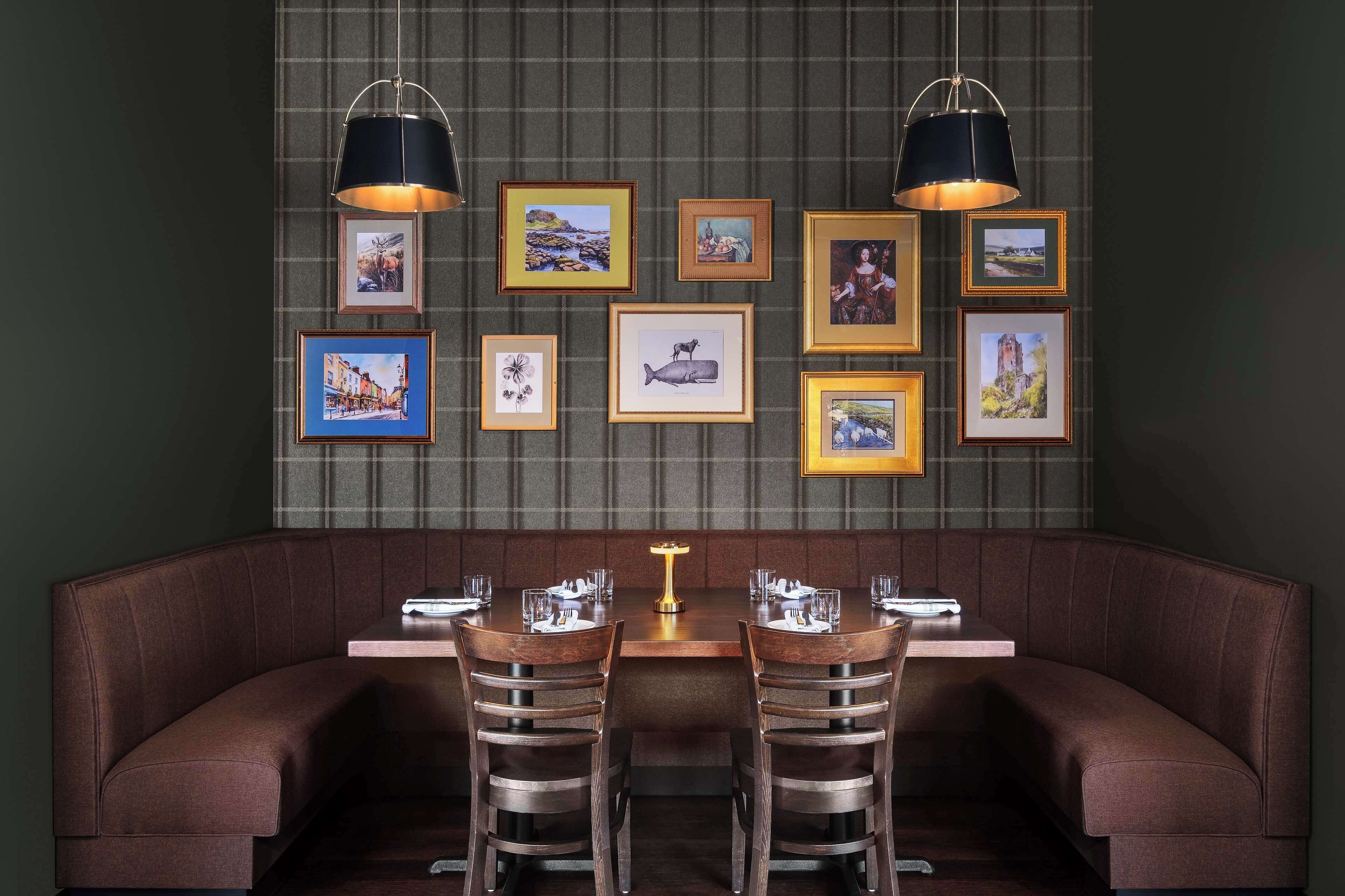 A restaurant with a dark green wall, a brown cushioned bench, and a table with two chairs. There are framed pictures on the wall above the bench.