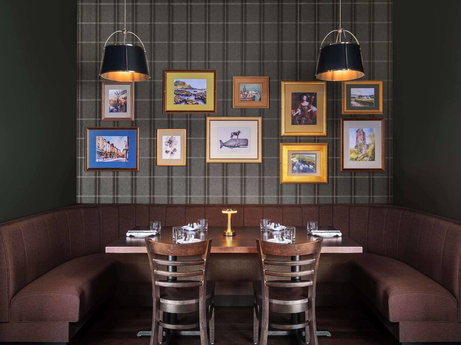 Un restaurant avec un mur vert foncé, un banc marron rembourré et une table avec deux chaises. Des tableaux encadrés sont accrochés au mur, au-dessus du banc.