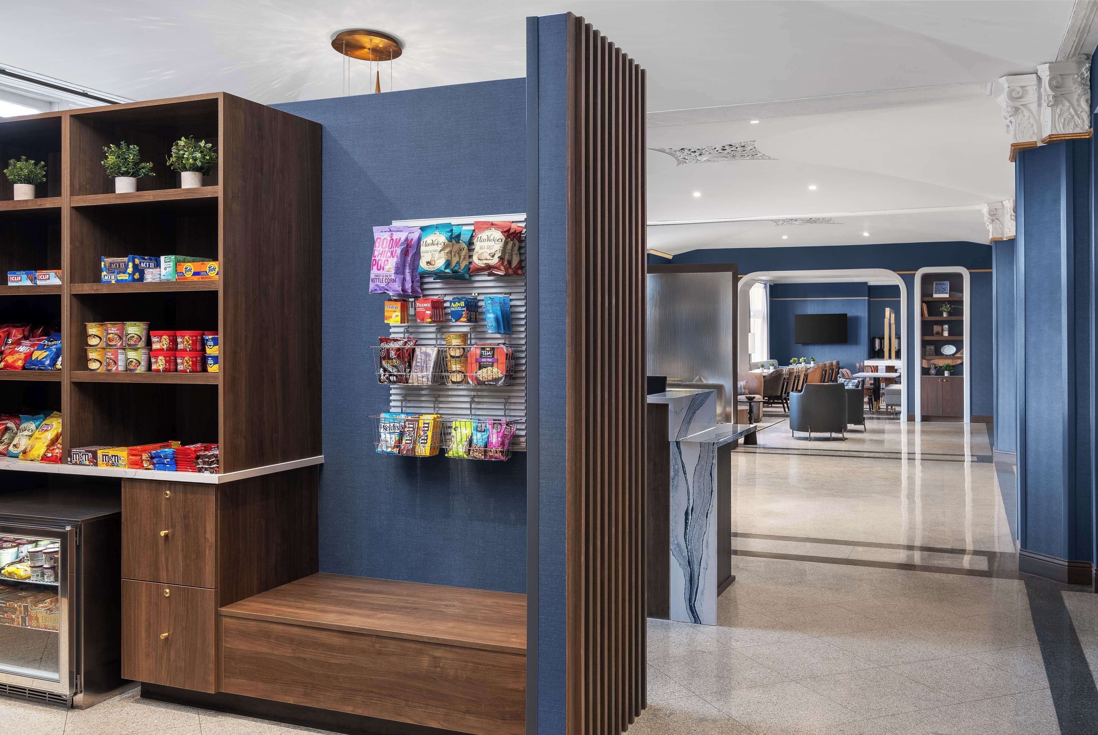 Un hall d’hôtel moderne avec un comptoir d’accueil en bois, un bar proposant divers produits emballés et un espace salon agrémenté d’un mur bleu et d’un sol en marbre.