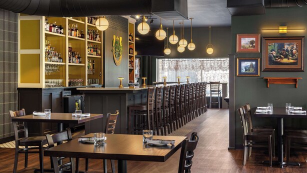 Un restaurant doté d’un long comptoir, de plusieurs tables et de chaises. On y trouve des étagères remplies de bouteilles et de verres, ainsi qu’un espace bar.