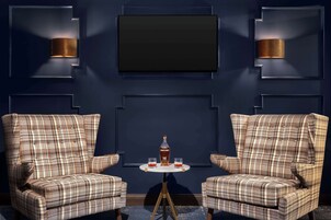 Una habitación azul oscuro con dos sillones de cuadros, una mesita redonda con una botella y vasos, y dos lámparas de pared.