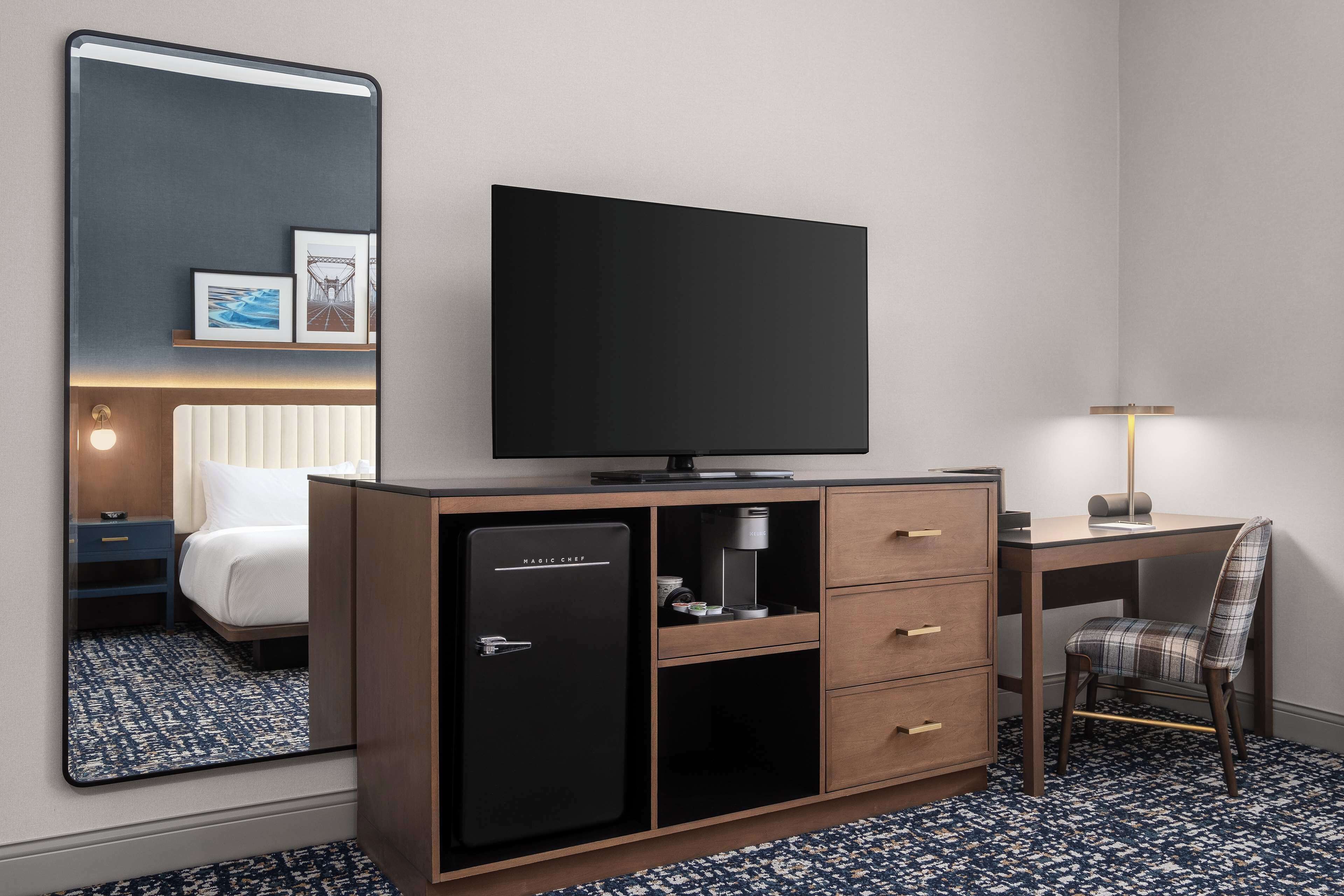 Habitación de hotel con un espejo grande, televisor de pantalla plana, minibar, escritorio con lámpara y una silla a cuadros.