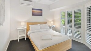 A bedroom with a bed, white nightstands, a ceiling fan, a painting, and a window with blinds. - Seaside Home- Direct beach access & pet friendly (Noosa)