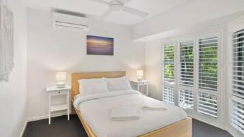 A bedroom with a bed, white nightstands, a ceiling fan, a painting, and a window with blinds.