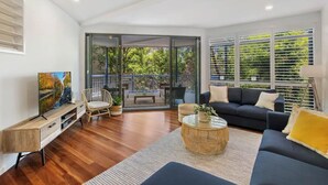 A modern living room with a TV, sofa, and coffee table. There is a sliding glass door leading to a balcony with outdoor furniture. - Seaside Home- Direct beach access & pet friendly (Noosa)