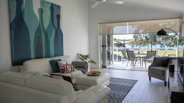 Ein modernes Wohnzimmer mit einem weißen Sofa, einem Couchtisch und einem großen abstrakten Gemälde an der Wand. Eine Glastür ermöglicht den Zugang zu einer Außenpatio-Terrasse mit Stühlen und einem Tisch.