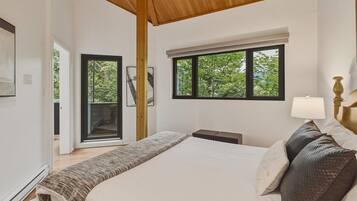 A bedroom with a large bed, a wooden ceiling, a window with a view of trees, and a door leading to an outdoor area.