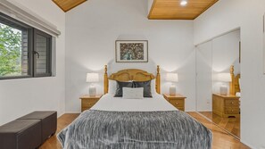 Un dormitorio con cabecera de madera, dos mesitas de noche con lámparas, una cama grande con colcha estampada gris, una ventana con persianas y un cuadro en la pared.