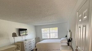 Un dormitorio con cama, escritorio, silla, cómoda, televisor y ventana.