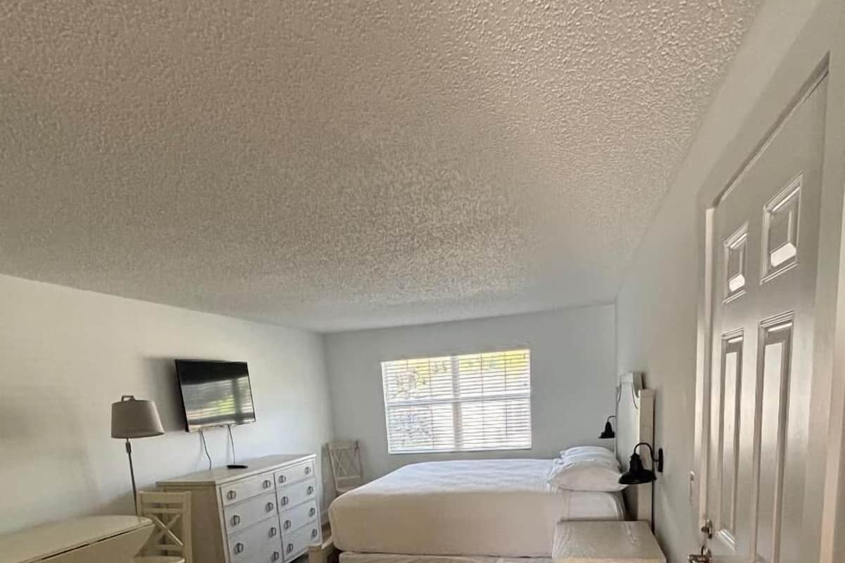 A bedroom with a bed, a desk, a chair, a dresser, a TV, and a window.